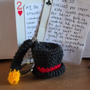 Llavero tejido a crochet en forma de sombrero de mago negro con cinta roja, apoyado sobre dos tarjetas numeradas de la edición limitada de Magia Sobre Ruedas